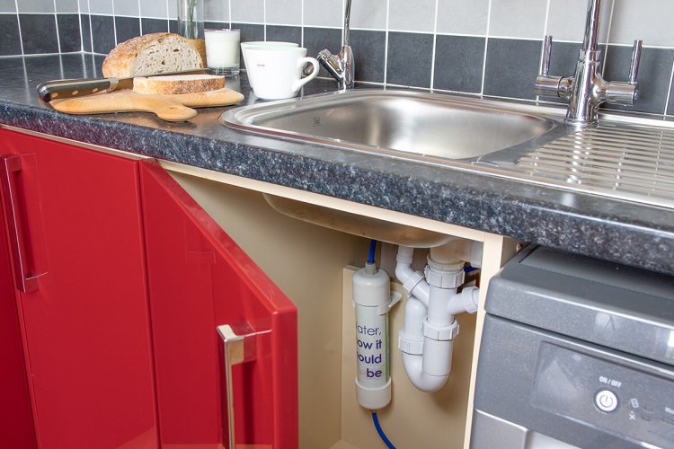 picture of a kitchen sink, board with bread, teacups, corner of a washing machine and red appliances with under sink water filter